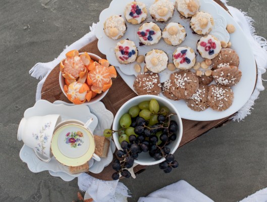 On the top tray: Vanilla Bean cakes filled with Blueberry Pomegranate Jam and topped with Coconut Cream Glaze, toasted coconut & macadamias, sugared lavender and rose petals. On the bottom tray, Spice Madeleines sprinkled with vanilla pearls. On the dessert plates under the tea cups: Lavender Lemon Shortbread.